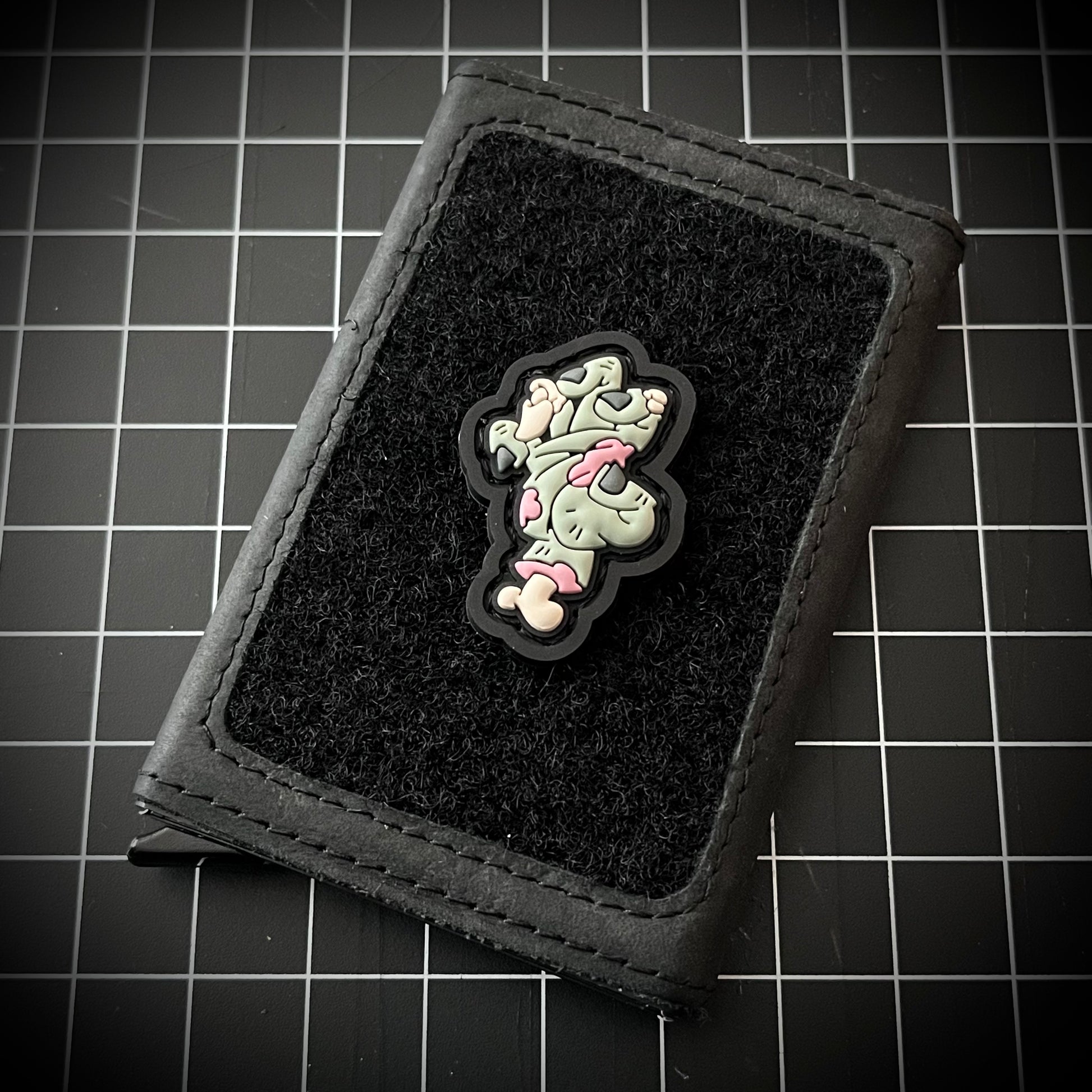 Zombie Hand RE PVC ranger eye morale patch of a green zombie hand with an exposed ring finger bone and wrist bone on a velcro wallet with a grid background.