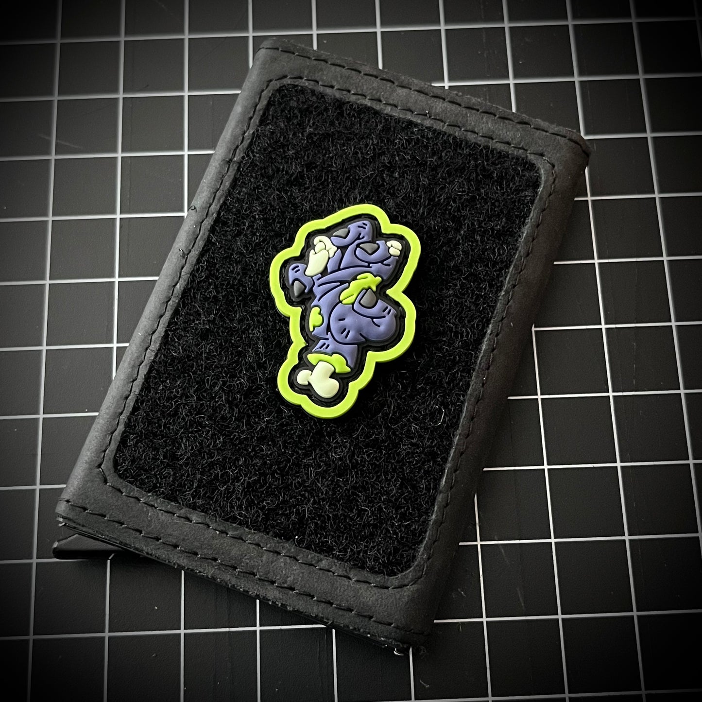 Zombie Hand RE - Toxic PVC ranger eye morale patch of a green and purple zombie hand with exposed bones on a velcro wallet with a grid background.